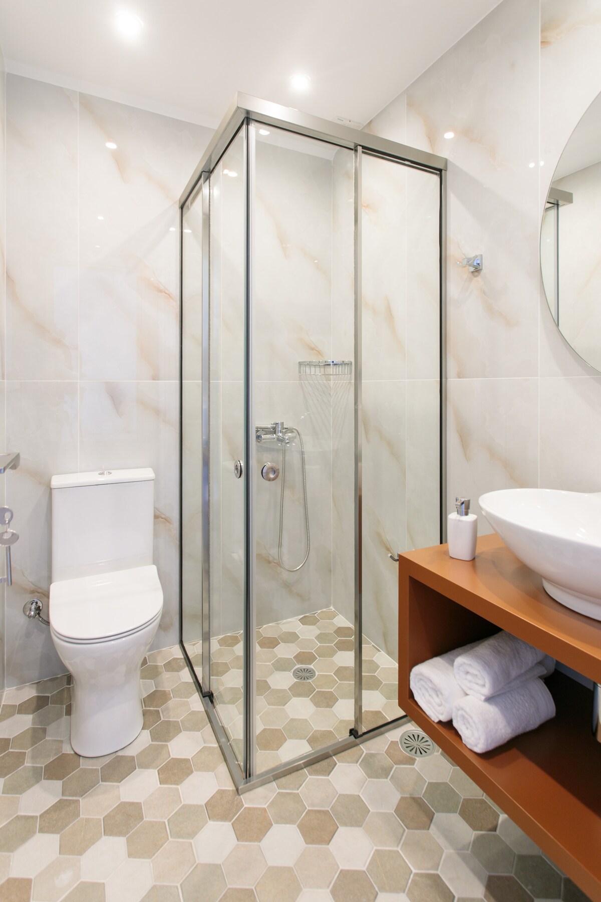 A modern bathroom space features a walk-in shower enclosed by glass doors. The room includes a sleek toilet and a wooden shelf with neatly arranged towels. Decorative hexagonal tiles cover the floor, while light-colored walls enhance the brightness of the area.