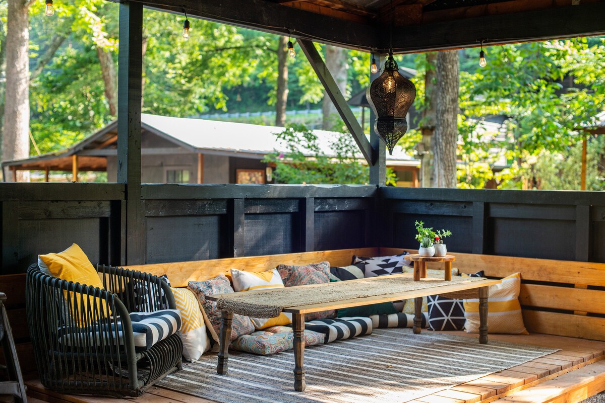 An outdoor seating area is presented with a rustic wooden table and an array of patterned cushions. String lights hang overhead, illuminating the space, while surrounding trees enhance the natural ambiance. The backdrop includes a glimpse of additional cabins in the distance.