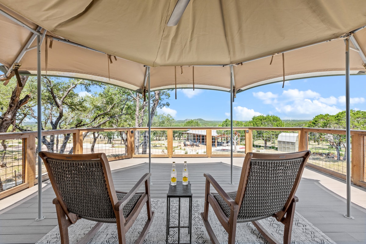A spacious deck is shown with two wooden rocking chairs placed opposite each other. A small table sits between them, holding two bottles. A panoramic view of the surrounding landscape and trees is visible through the tent-like covering.
