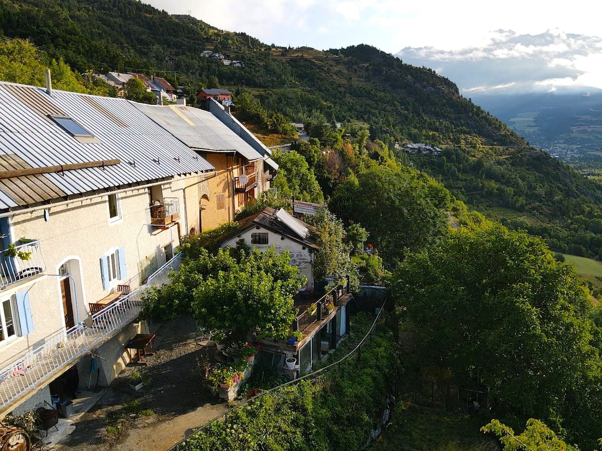 A panoramic view of a charming hillside property is captured, displaying multiple levels of the building. Lush greenery surrounds the home, with trees and plants providing a natural frame. A balcony extends from the upper floor, offering a serene outdoor space with scenic mountain vistas.