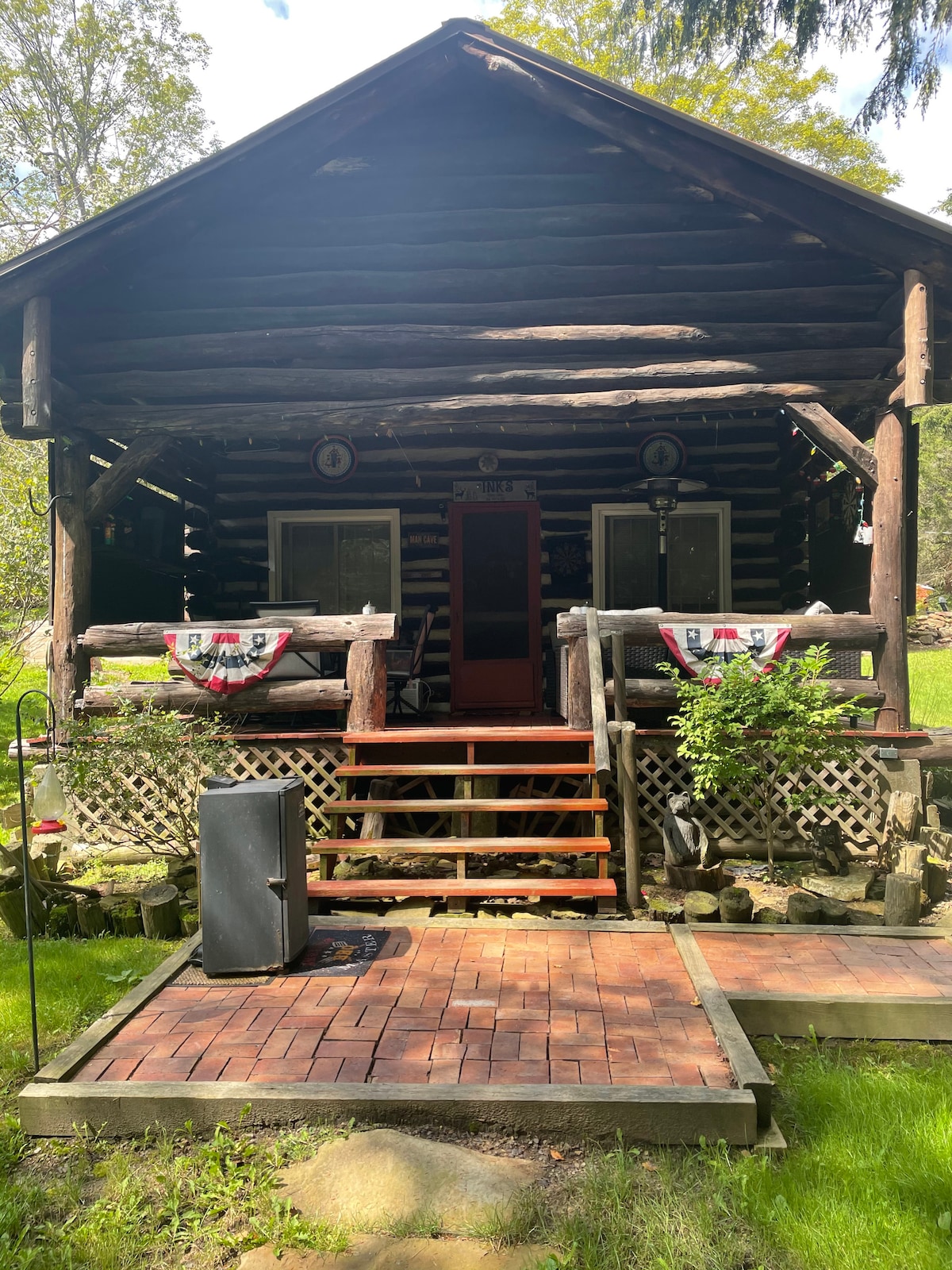 The log cabin exterior features a welcoming front porch with steps leading to the entrance. The porch is adorned with decorative banners and surrounded by greenery. The cabin's rustic charm is complemented by a wooden railing and a stone border along the garden area.