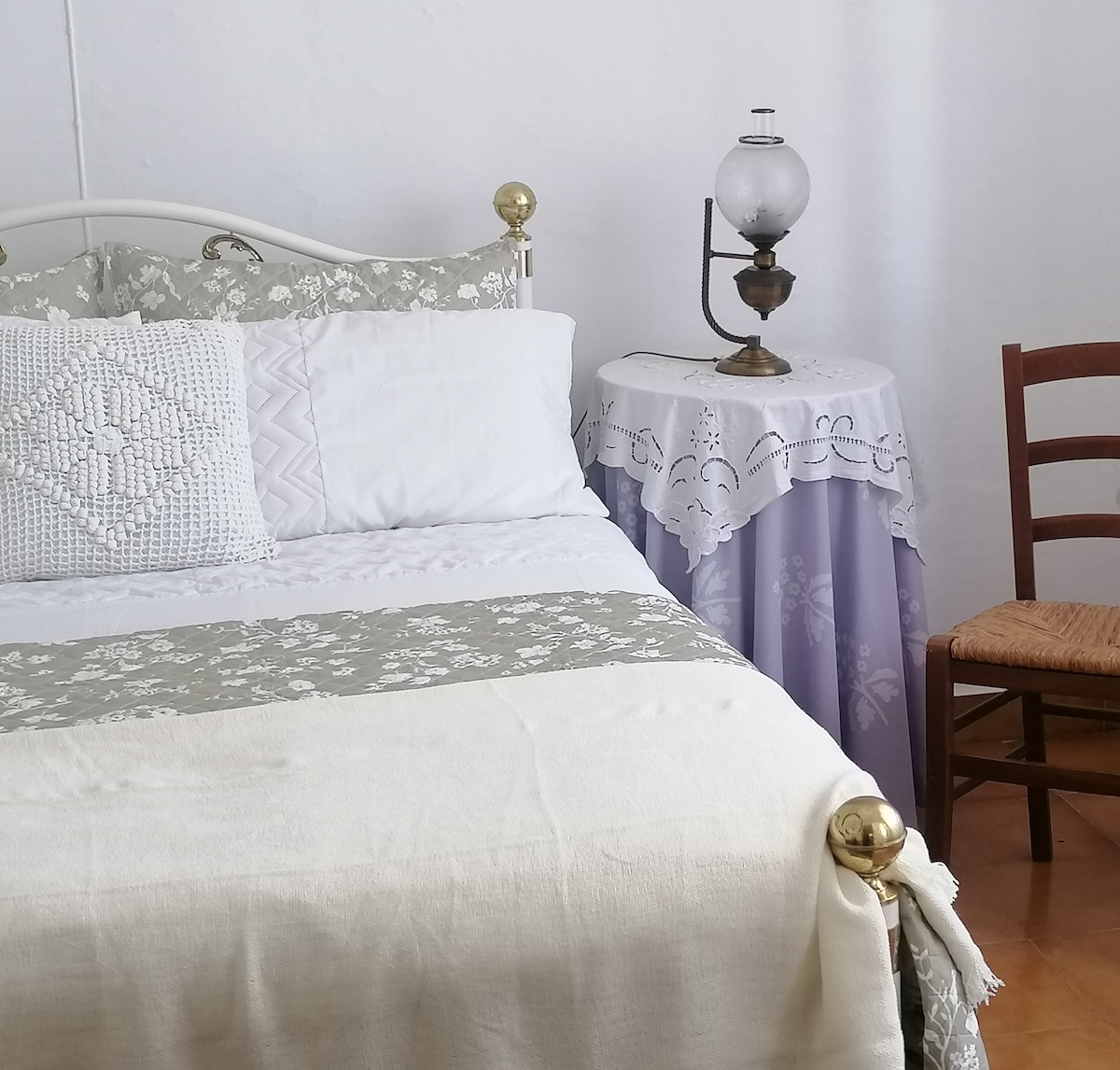 A quaint bedroom features a classic bed adorned with a light-colored blanket and decorative pillows. A small bedside table with a lace cover holds an antique lamp. A wooden chair adds to the room's charm, with warm light reflecting off the white walls.