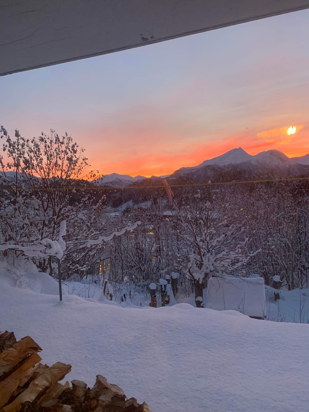 A winter landscape features snow-covered trees and mountains in the background. The sky is adorned with hues of orange and pink, reflecting a tranquil sunset. Soft snow blankets the ground, creating a serene atmosphere in a secluded setting.