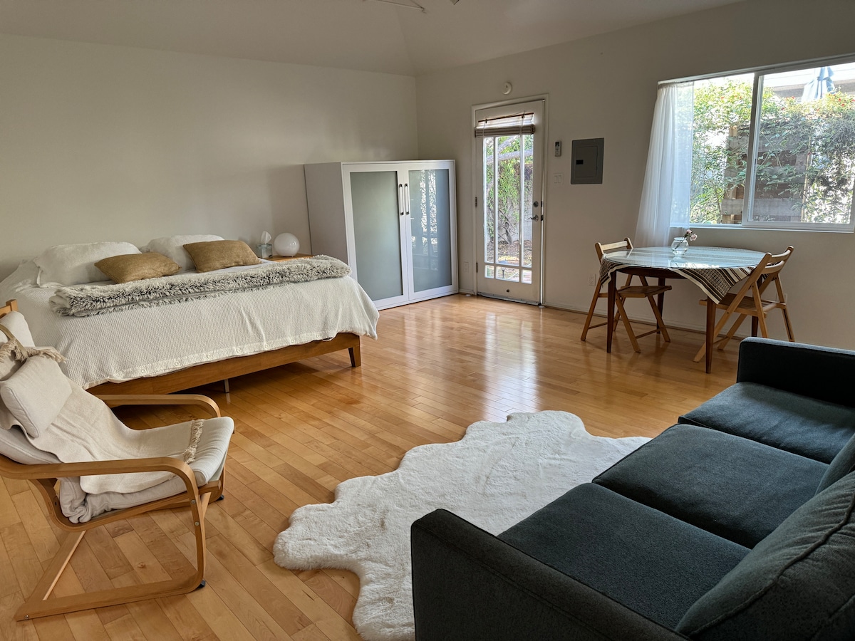 A spacious studio is presented with wooden floors and ample natural light coming through the windows. A king size bed with decorative pillows is positioned next to a sliding glass door leading to a garden. A small dining table with chairs is situated beside a cozy seating area.