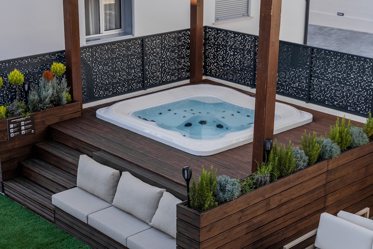 An outdoor lounge area is highlighted by a heated jacuzzi, surrounded by wooden decking and lush greenery. Comfortable seating is arranged nearby, offering a relaxing space with decorative plants and a modern fenced design showcasing intricate patterns.