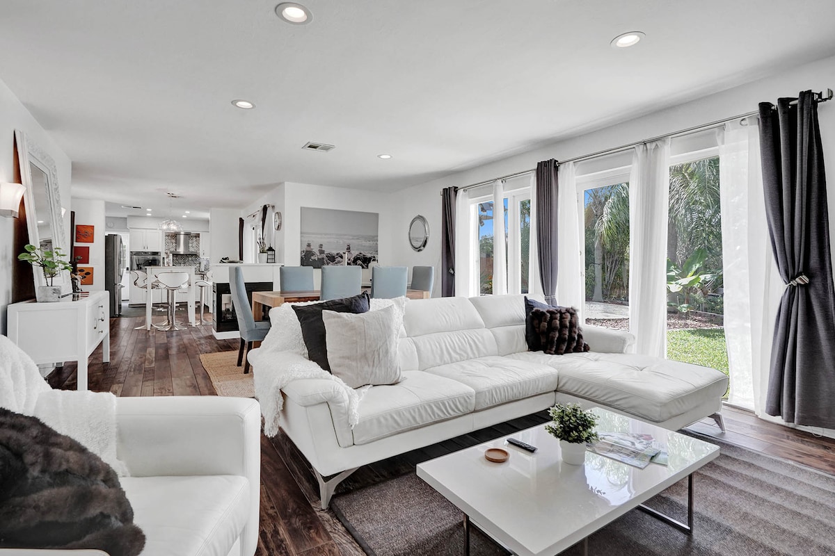 A bright living area is furnished with a spacious white sectional sofa positioned on a light rug. Large windows allow natural light, revealing views of the outdoor space. Dining and kitchen areas are visible in the background, enhancing the sense of openness.