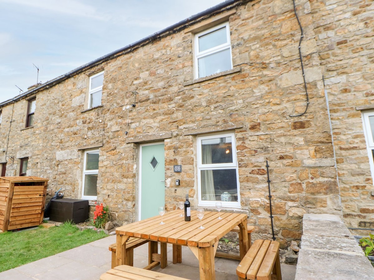 The exterior of a traditional Yorkshire stone cottage is depicted, featuring a light green door and two upper windows. A wooden picnic table with benches sits on a paved area, accompanied by a bottle of wine. Lush grass and flowers add a touch of greenery.
