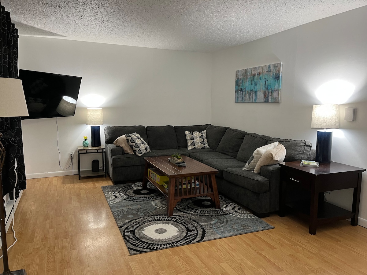A comfortable living area features a large gray sectional sofa arranged around a wooden coffee table. Two stylish lamps provide soft lighting, and a patterned rug adds texture. A modern piece of wall art complements the space, enhancing the inviting atmosphere.