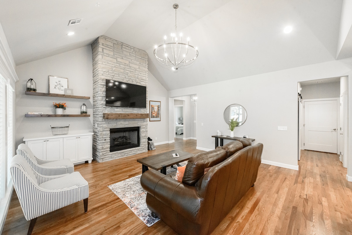 A spacious living area features a large stone fireplace under a soaring ceiling. Comfortable seating is provided by a leather sofa and striped chairs. A wooden coffee table sits centrally, with a subtle area rug enhancing the warm wood flooring. Neutral tones dominate the decor.