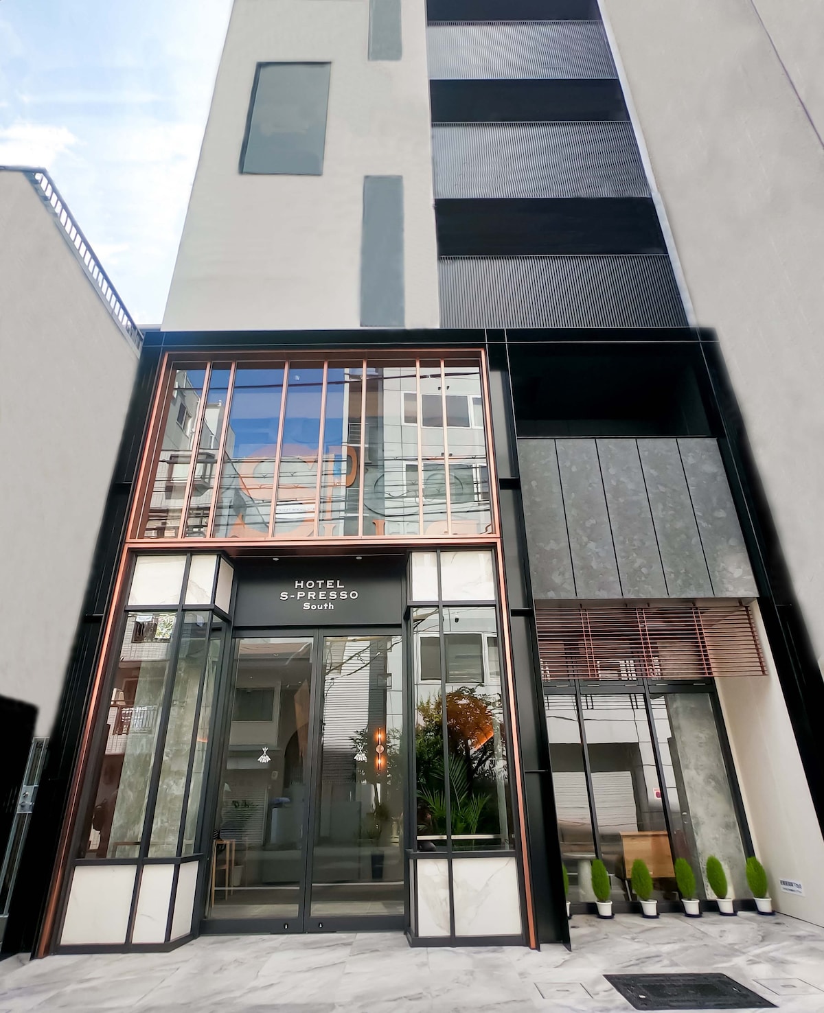 The contemporary building features a modern façade with large glass windows and an inviting entrance. Decorative plants are arranged in neat rows outside, complementing the sleek architectural lines of the structure.