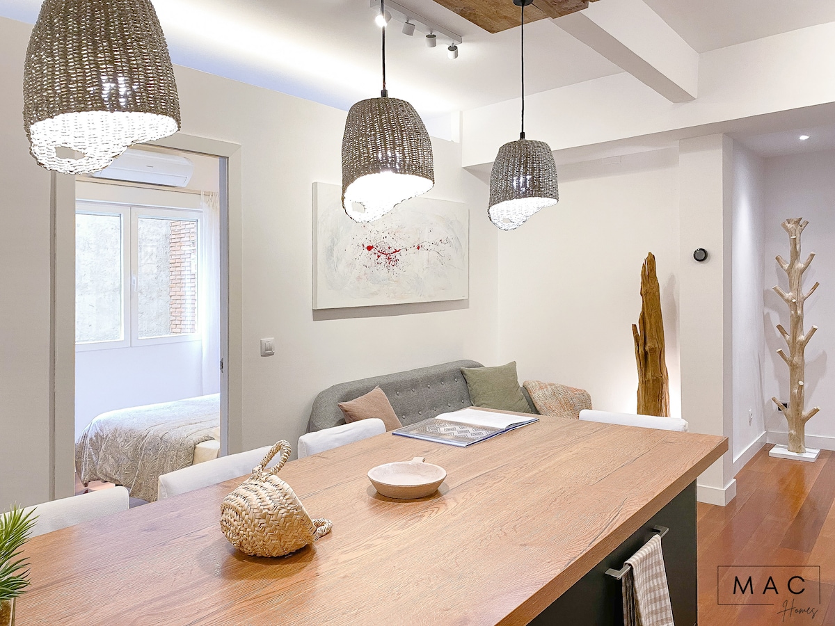 A modern dining area features a large wooden table with a round white plate and a decorative basket. Three woven pendant lights hang above, illuminating the space. In the background, a comfortable sofa is positioned near a large window, with a glimpse of a bedroom visible through the doorway.