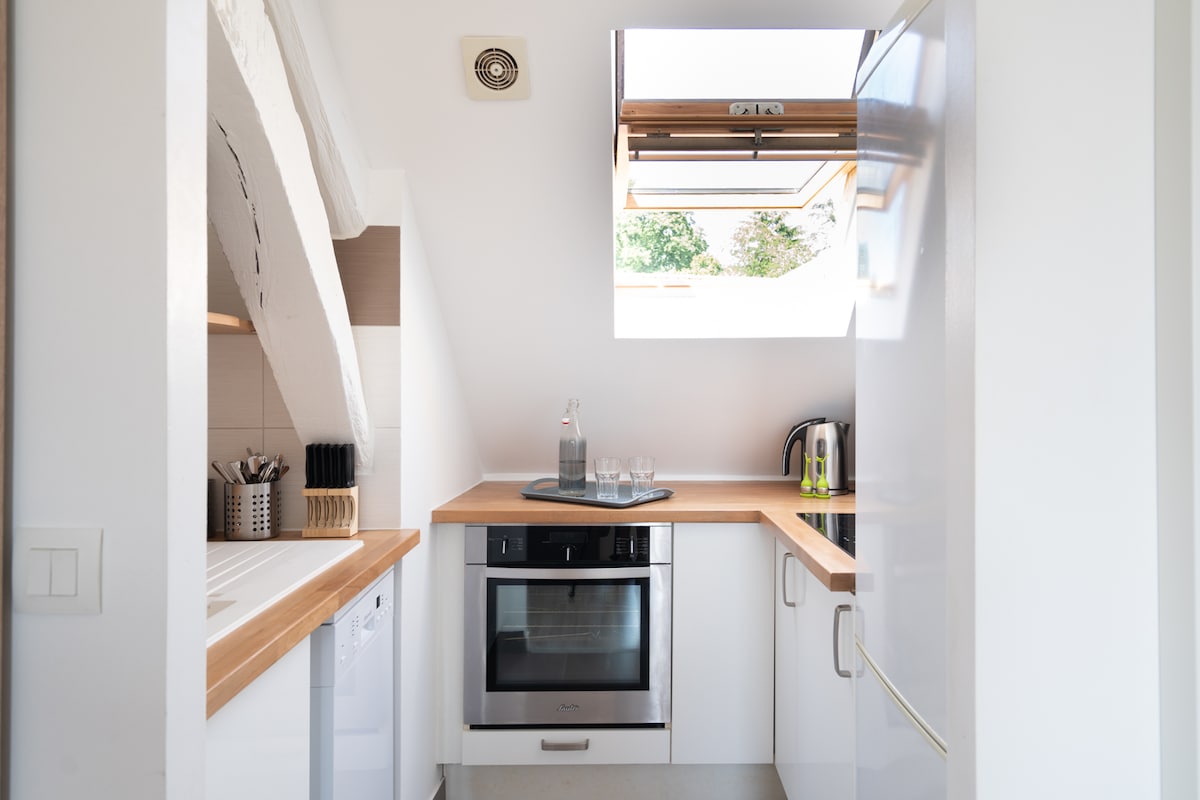 The kitchen features a sleek design with white cabinetry and a wooden countertop. Essential appliances, including an oven and a refrigerator, are positioned for ease of use. Natural light enters through a skylight, enhancing the bright and clean atmosphere of the space.