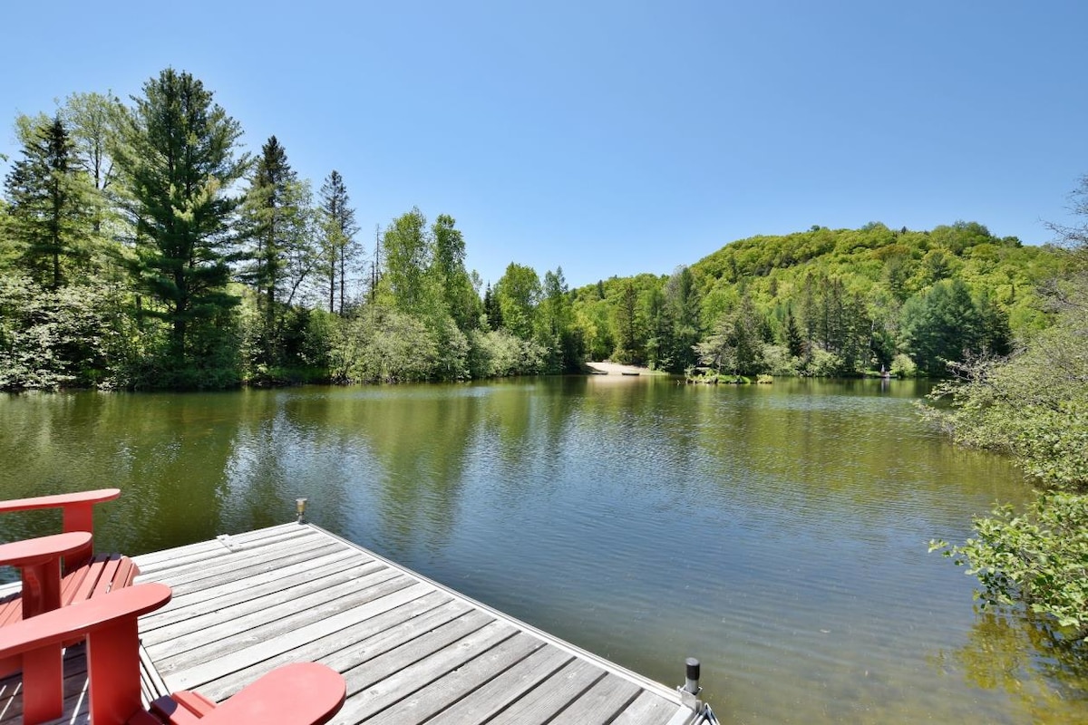 A private dock extends over a tranquil lake, surrounded by lush greenery and trees. Bright red Adirondack chairs are positioned invitingly on the dock, offering a perfect spot to enjoy the peaceful water views and natural scenery.
