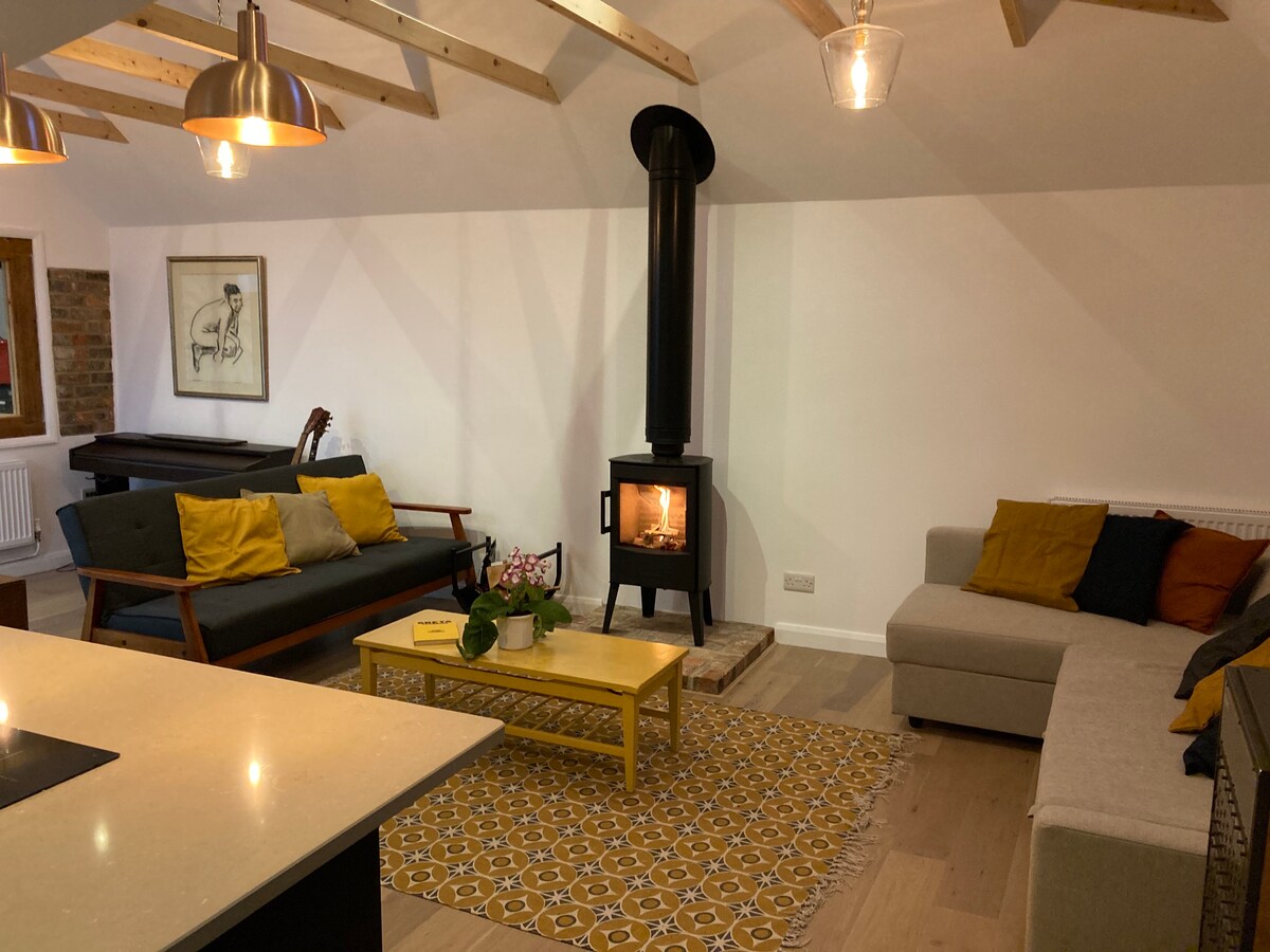 An open plan living area is presented with a mix of seating options, including a dark sofa and a light-colored sectional. A log burner stands prominently, providing warmth. Soft lighting emanates from pendant fixtures above, while a patterned rug adds texture to the wood flooring.