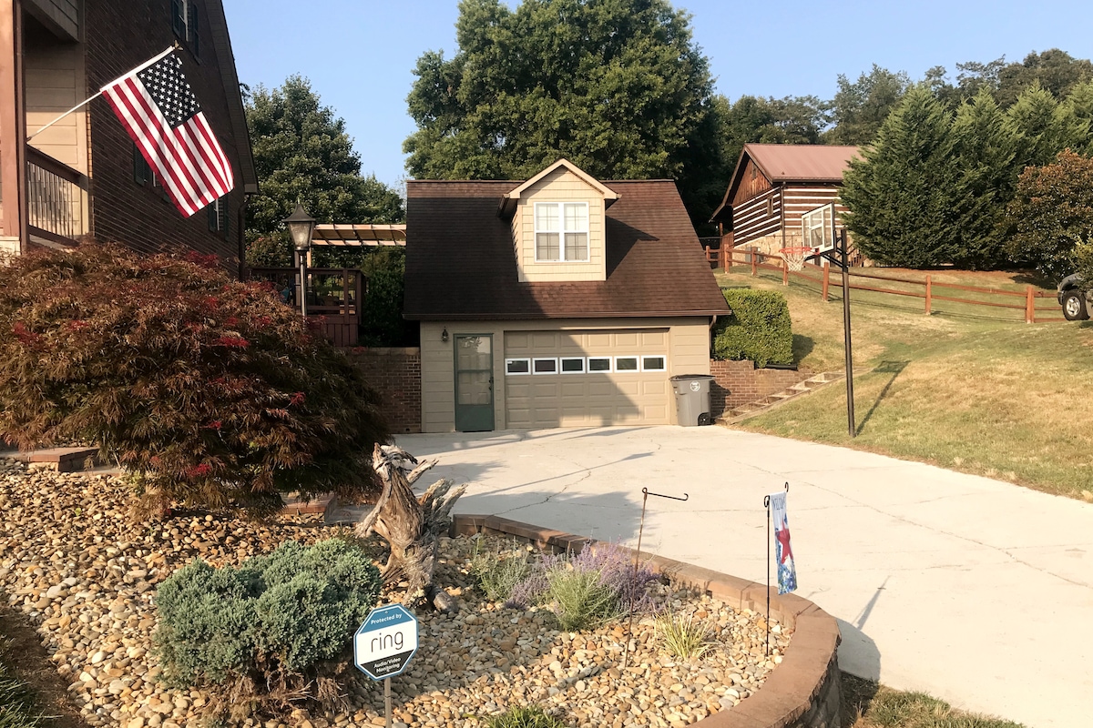 A peaceful exterior view showcases a well-maintained driveway leading to a spacious garage. The property features a charming loft above the garage, surrounded by landscaped gardens with stones and plants. An American flag is prominently displayed, enhancing the welcoming ambiance.