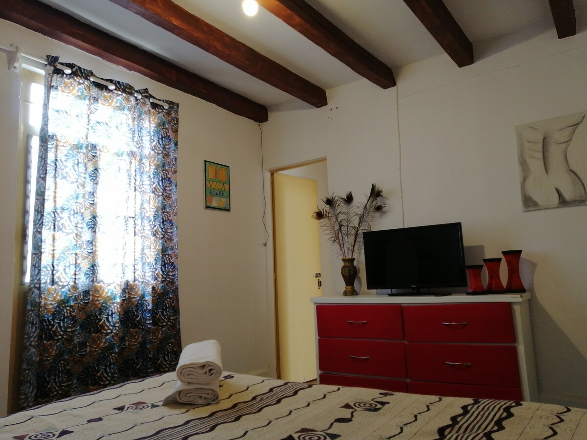 A brightly lit bedroom features wooden beams across the ceiling and a colorful patterned curtain filtering natural light. A large bed is adorned with a textured cover, and a television is mounted on a red dresser, complemented by decorative items and a vase.