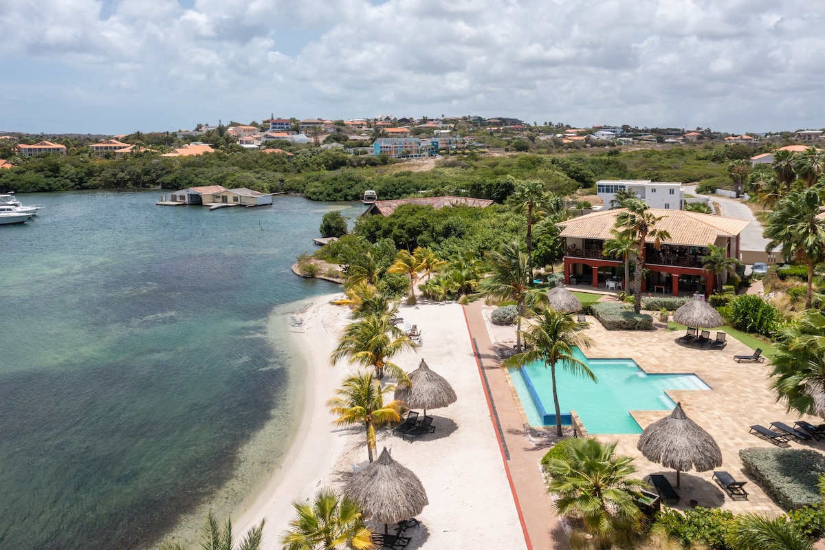 An aerial view captures the serene landscape featuring an infinity pool adjacent to a private beach. Lush palm trees provide shade over thatched umbrellas, while the tranquil waters of the Spanish Water reflect the clouds above. Nearby, villas dot the hillside, creating a peaceful atmosphere.