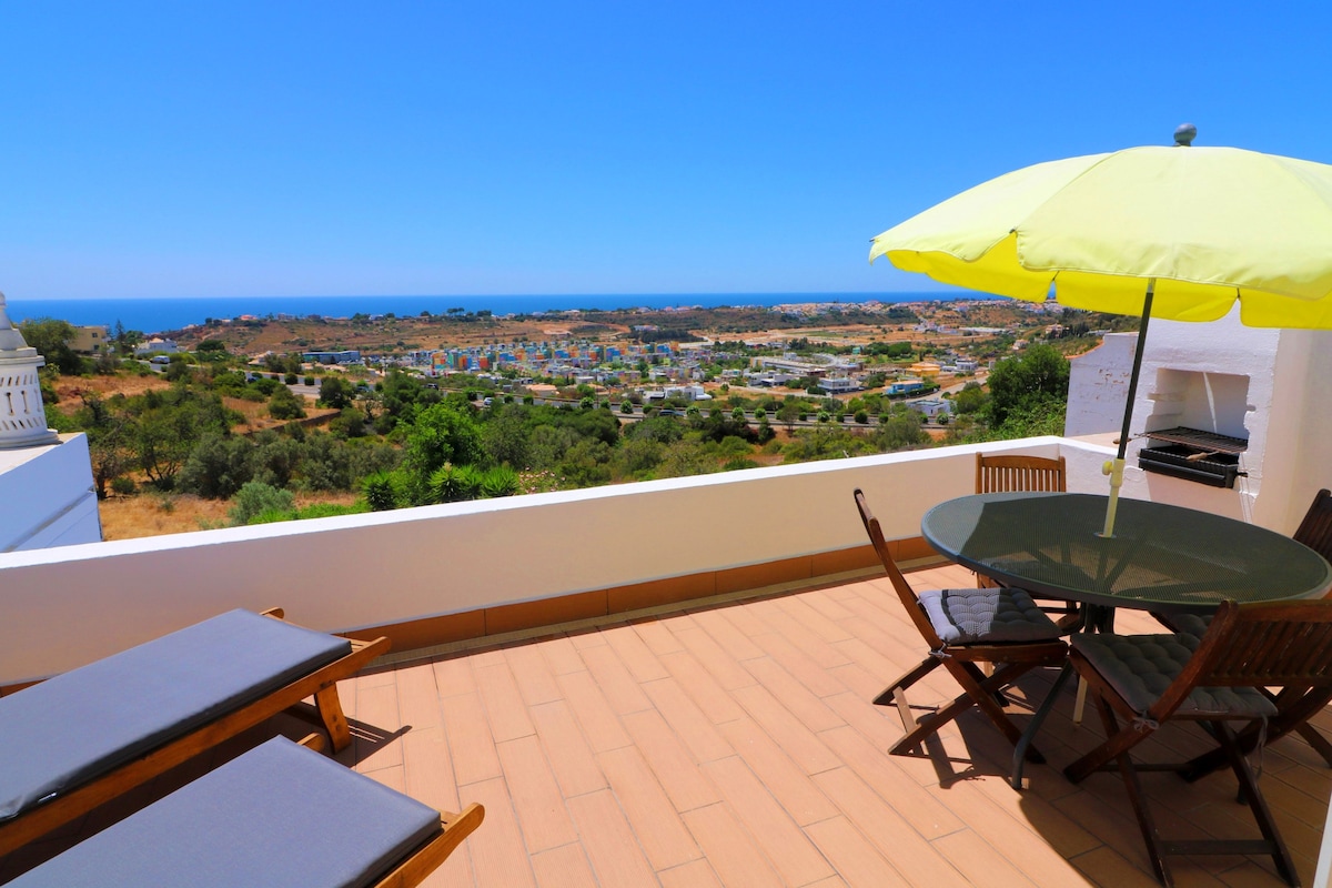 A spacious balcony offers panoramic views of the surrounding landscape, including the sea in the distance. Sun loungers are positioned beside a round table shaded by a bright yellow umbrella. A charcoal grill is visible, enhancing the outdoor dining experience.