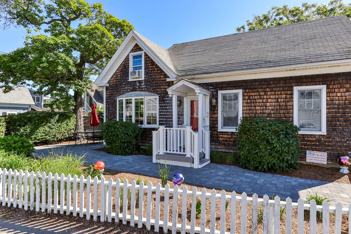The exterior showcases a charming house with a shingled facade and a welcoming entryway. A white picket fence surrounds the landscaped yard, which features native plants and decorative elements. Large windows allow for natural light, and a patio with an umbrella is visible to the side.