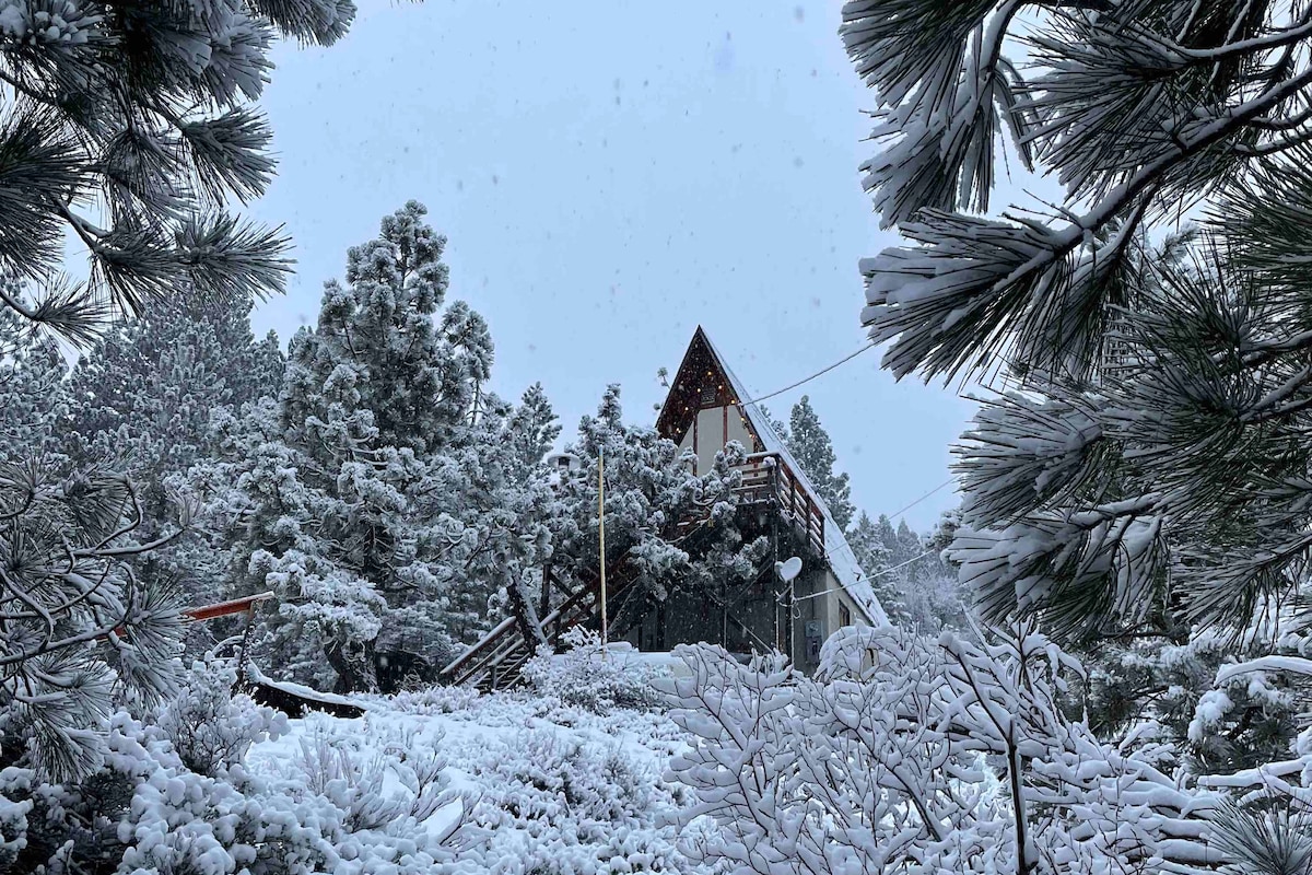 The A-frame chalet is surrounded by a blanket of snow, with evergreens framing the scene. The structure showcases a sloped roof and a front deck, partially obscured by snow-laden branches. The atmosphere is quiet and serene, characteristic of a winter landscape.