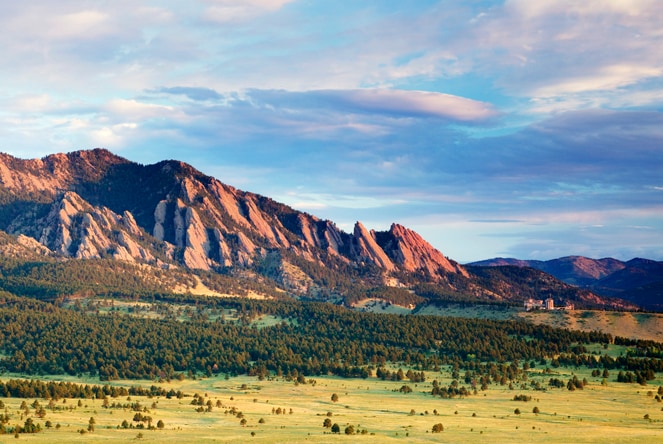 An expansive view of a mountain range stretches across the horizon. The rugged rock formations are bathed in soft light, contrasting with the lush green landscape below. Fluffy clouds drift overhead, creating a serene atmosphere that reflects the natural beauty of the area.