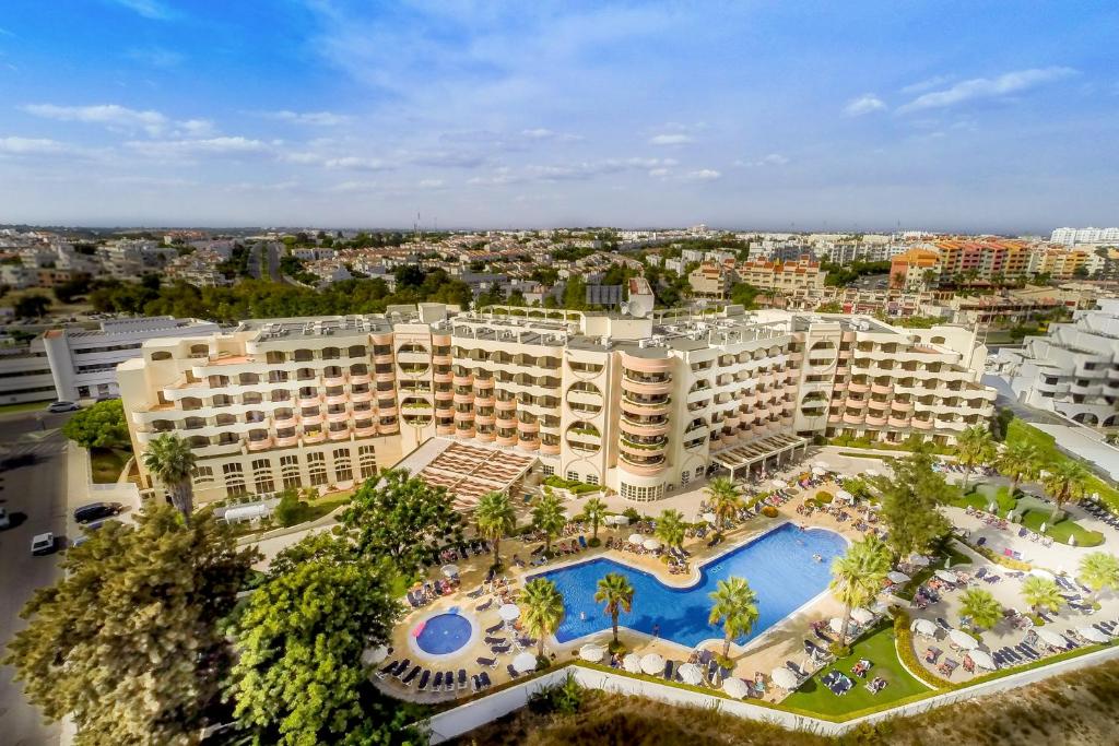 An aerial view of a modern hotel showcases its multiple levels and expansive outdoor pool surrounded by palm trees. Sun loungers are arranged around the pool area, and the vibrant cityscape lies in the background, highlighting the hotel's spacious layout and inviting environment.