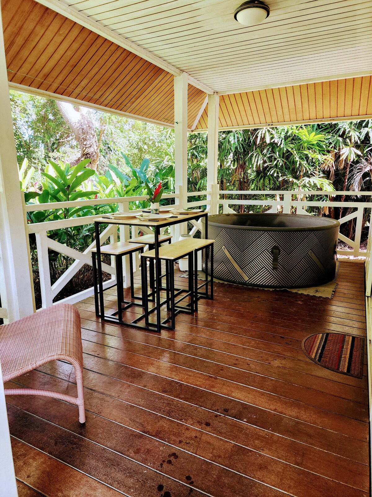 An outdoor terrace features a stylish hot tub and a set of simple dining furniture, including a table and stools. Lush greenery surrounds the area, enhancing the tranquil atmosphere, while warm wooden flooring adds to the inviting space.