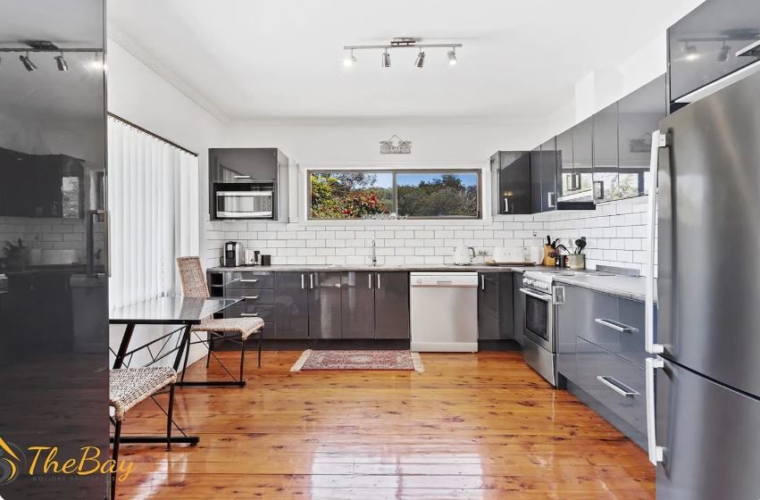 A modern kitchen is equipped with sleek gray cabinetry and a white subway tile backsplash. Stainless steel appliances are present, along with a dining table and two chairs, creating an inviting space for meal preparation and dining. Natural light fills the room through a large window.