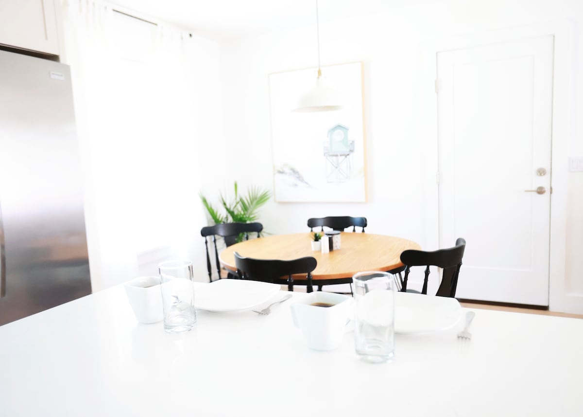 A bright dining area features a round wooden table surrounded by black chairs. The table is set with white dishes and glassware. A simple light fixture hangs above, while a green plant is visible in the background, enhancing the clean and airy atmosphere.