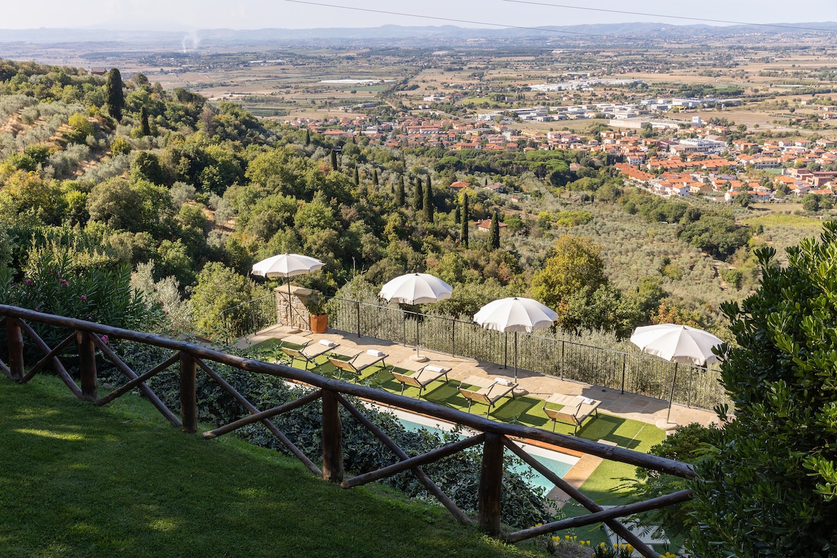 A view from a terrace overlooking a lush valley with rolling hills. Several umbrellas provide shade over outdoor seating areas, overlooking a pool framed by sunbeds. The area is surrounded by greenery, with distant hills and small towns visible under a clear sky.