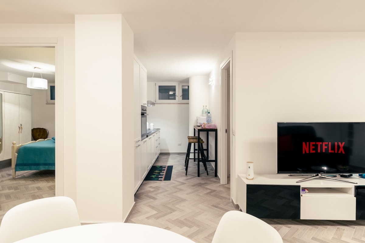 An open-plan living area is visible, featuring a modern television displaying the NETFLIX logo. A streamlined kitchen with white cabinetry is in the background, complemented by a small dining table and chairs. Ample natural light fills the space through several windows.