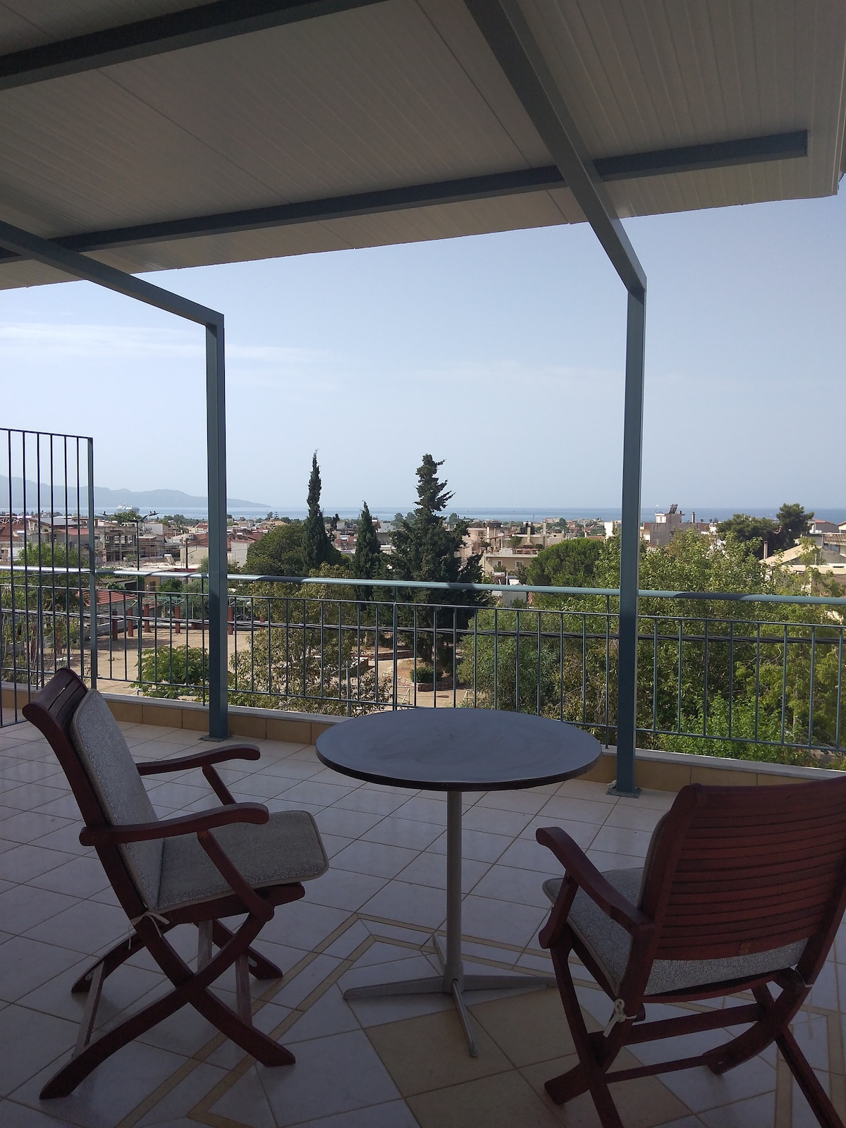 A balcony area features a round table paired with two comfortable chairs, offering a view of the surrounding trees and the distant coastline. The open space is framed by a modern roof structure, providing an inviting spot to relax outdoors.