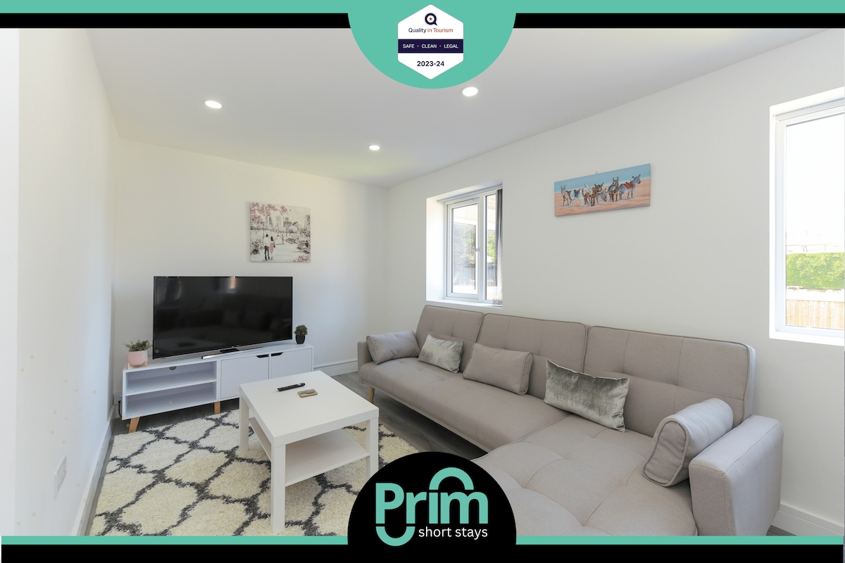 A modern living area is presented, featuring a spacious sectional sofa and a flat-screen TV on a minimalist console. Natural light fills the room through large windows, and a light-colored area rug adds warmth to the space.
