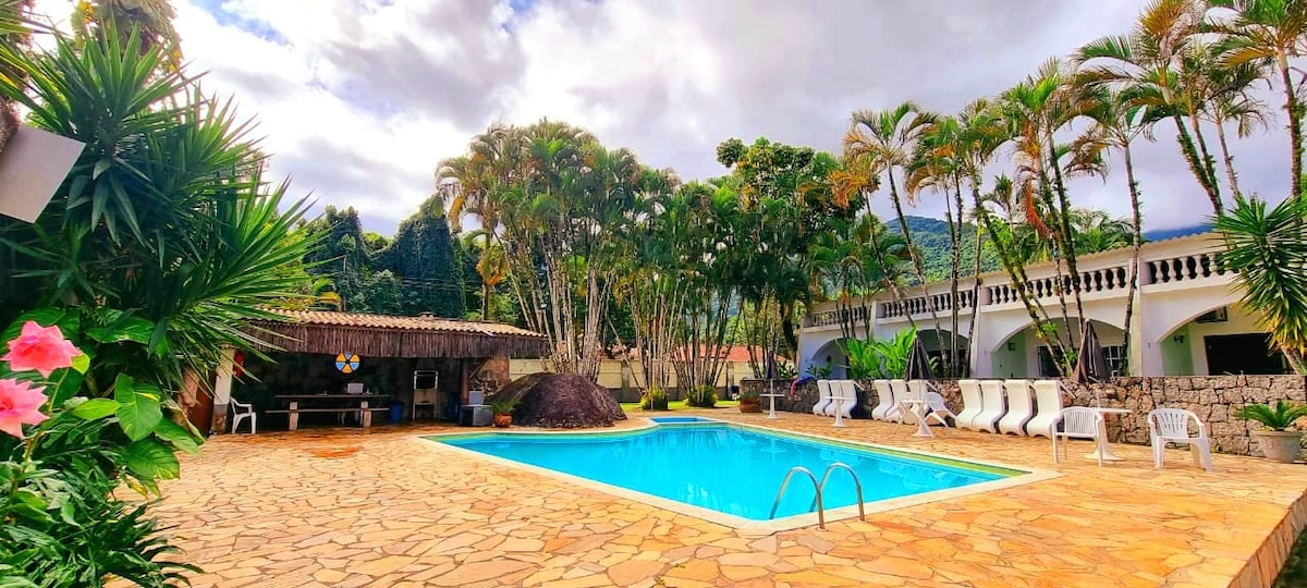 A serene outdoor space features a clear blue swimming pool surrounded by lush palm trees and tropical foliage. A natural rock formation is present nearby, and several lounge chairs are positioned along the poolside. A shaded gathering area is visible, offering a tranquil setting.