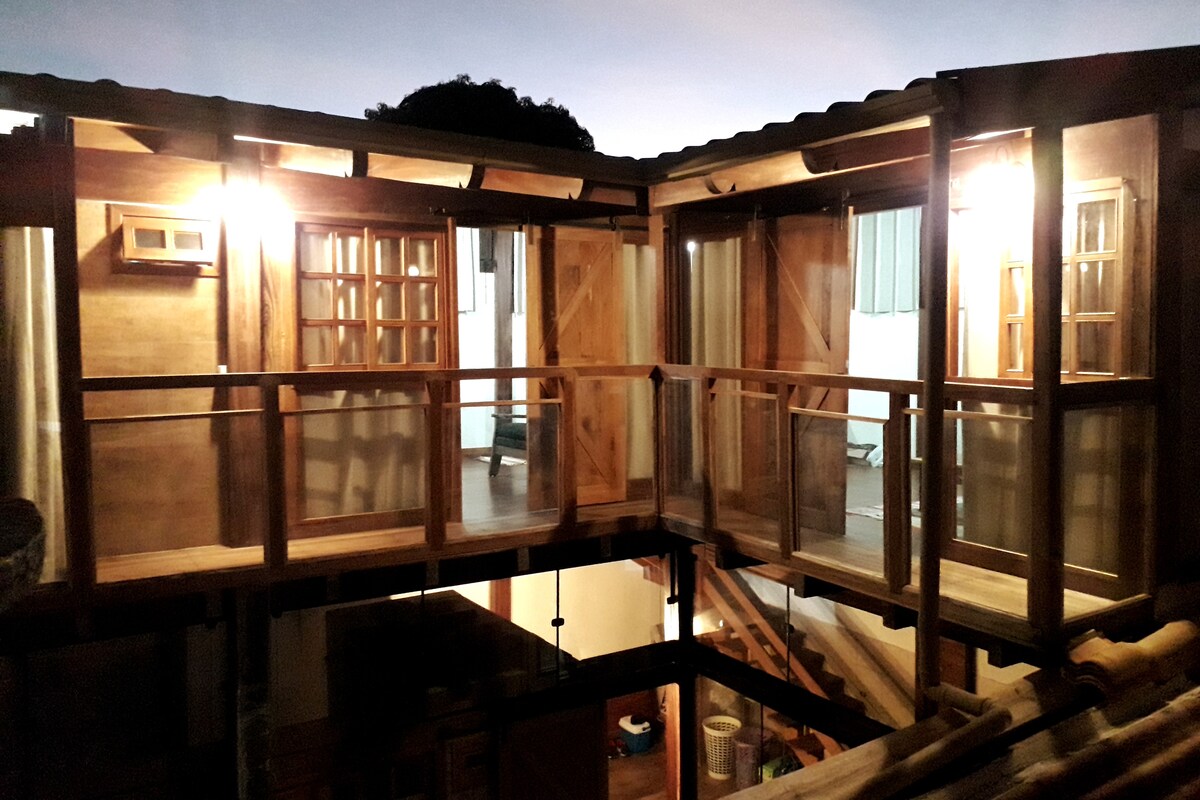 A wooden structure with large glass windows and doors is illustrated, allowing natural light to flow into the space. The design features a balcony area and a staircase leading to the floor below, enhancing accessibility and the connection between levels.