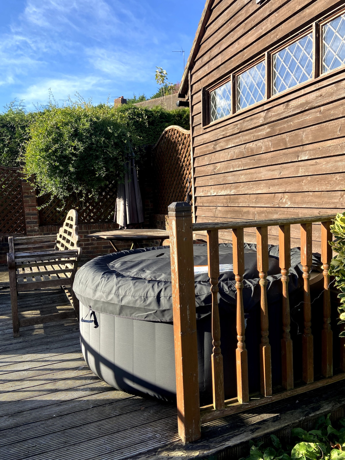 A private hot tub is situated on a wooden deck, surrounded by greenery and decorative lattice. Rustic wooden benches are positioned nearby, providing a comfortable space for relaxation. The exterior of the building features warm brown wood paneling and several window panes reflecting natural light.