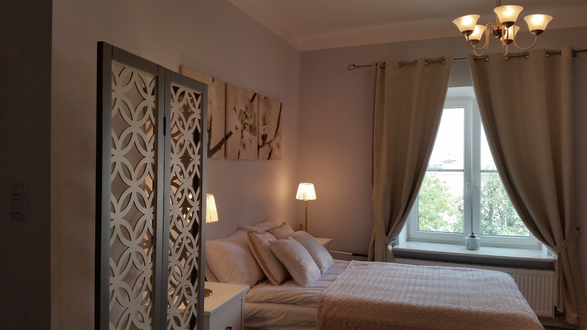 A sleeping area is highlighted by soft, neutral tones and elegant decor. A spacious bed is positioned under a window with light curtains, providing natural illumination. Two bedside lamps add a gentle glow, and a decorative screen offers a partition to enhance privacy.