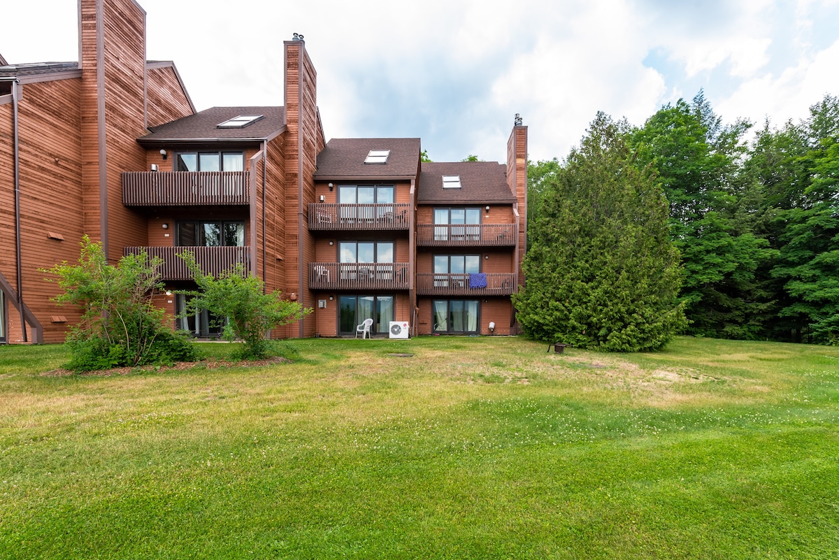 The exterior view features a multi-story wooden building surrounded by lush green grass. Multiple balconies provide access to outdoor spaces, while trees frame the backdrop, enhancing the natural setting.