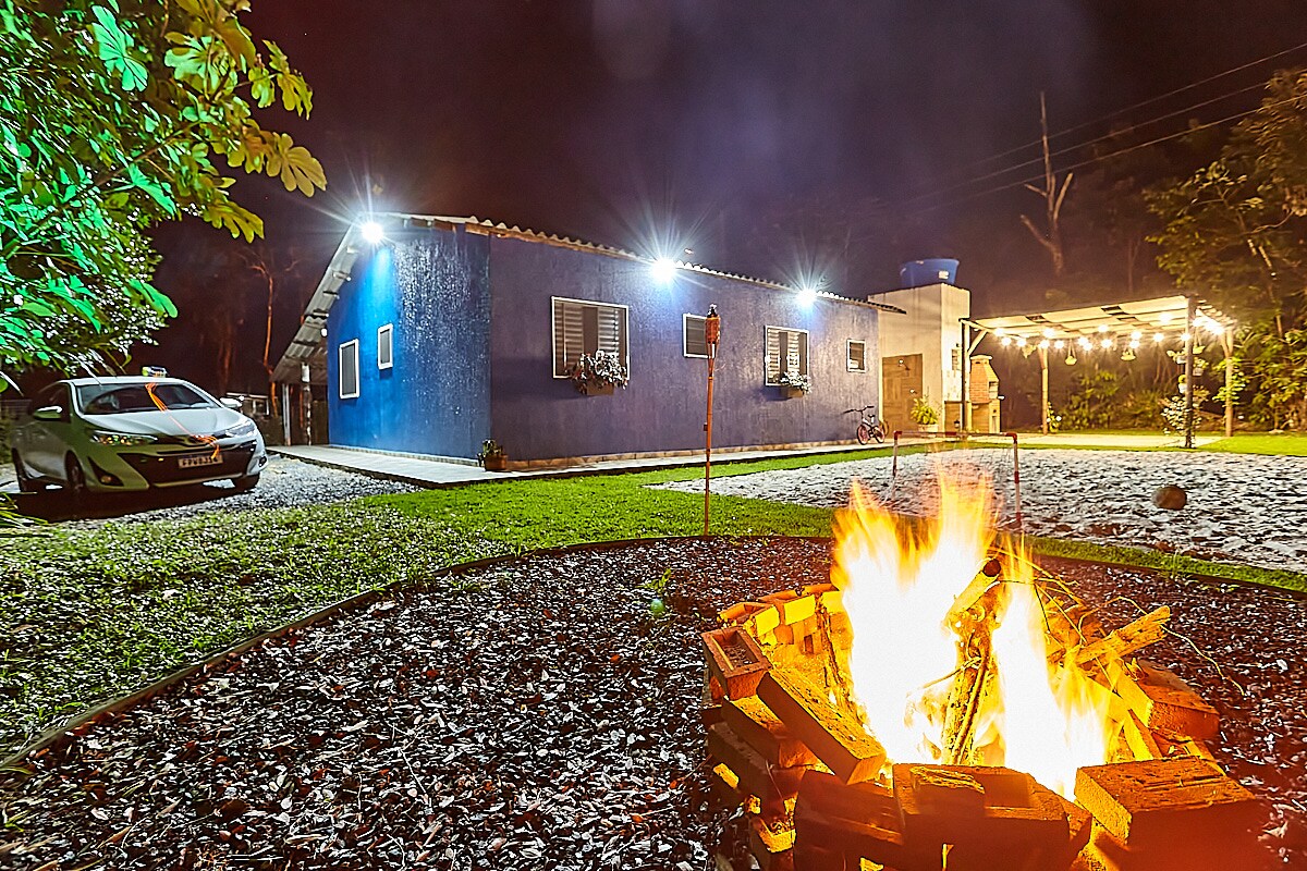 A welcoming outdoor space is illuminated at night by warm lights around a fire pit. A blue house is visible in the background, with a covered area to one side and a parked car in the driveway, surrounded by green grass and trees.