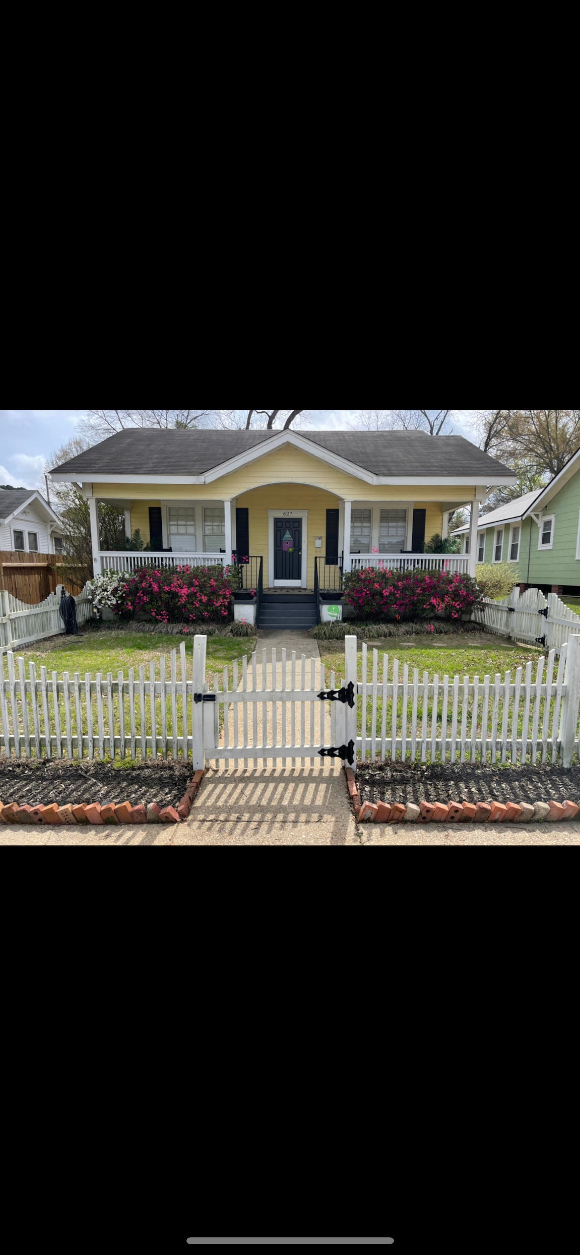 The charming cottage is framed by a white picket fence and features a welcoming front pathway. Colorful flowers bloom along the porch, which hosts two rocking chairs. A front door leads into the inviting home, complemented by lush green grass and a well-maintained landscape.