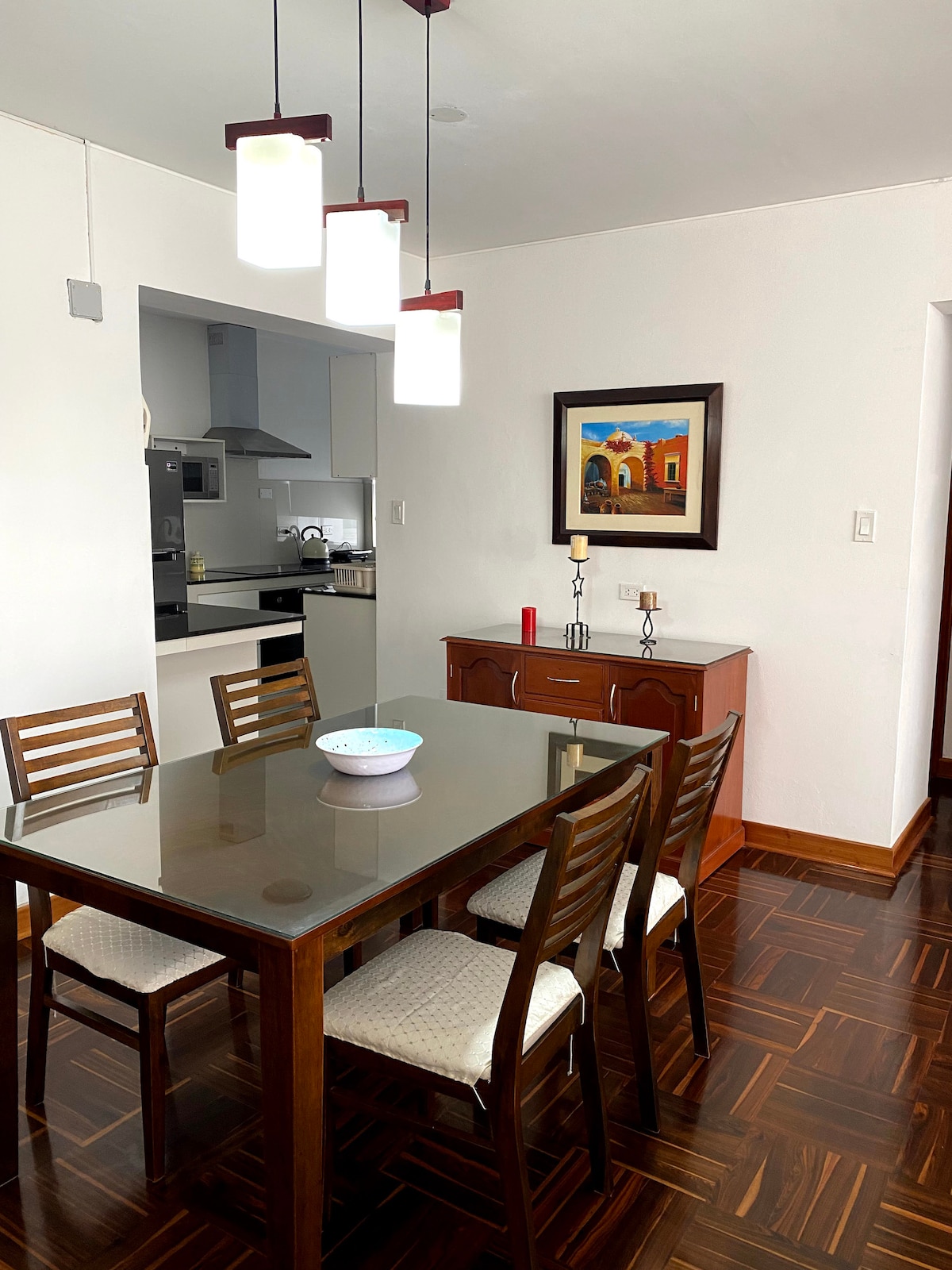 An inviting dining area is highlighted by a rectangular wooden table surrounded by six chairs. Above, three modern pendant lights provide ample illumination. A cabinet with decorative items complements the space, and a piece of artwork featuring a vibrant building adds a touch of color to the wall.