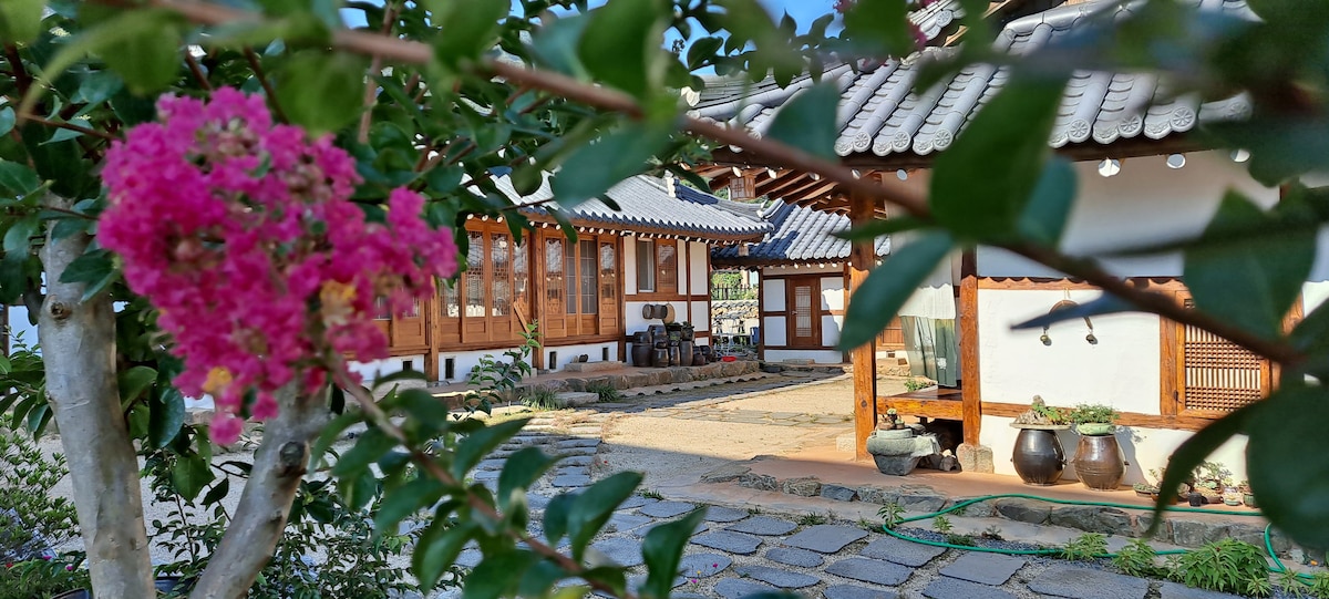 A serene view of a traditional Hanok is framed by vibrant pink flowers. The architecture features wooden accents and a distinctive tiled roof, with a stone pathway leading through the courtyard. Traditional ceramic pots and greenery are visible in the background.