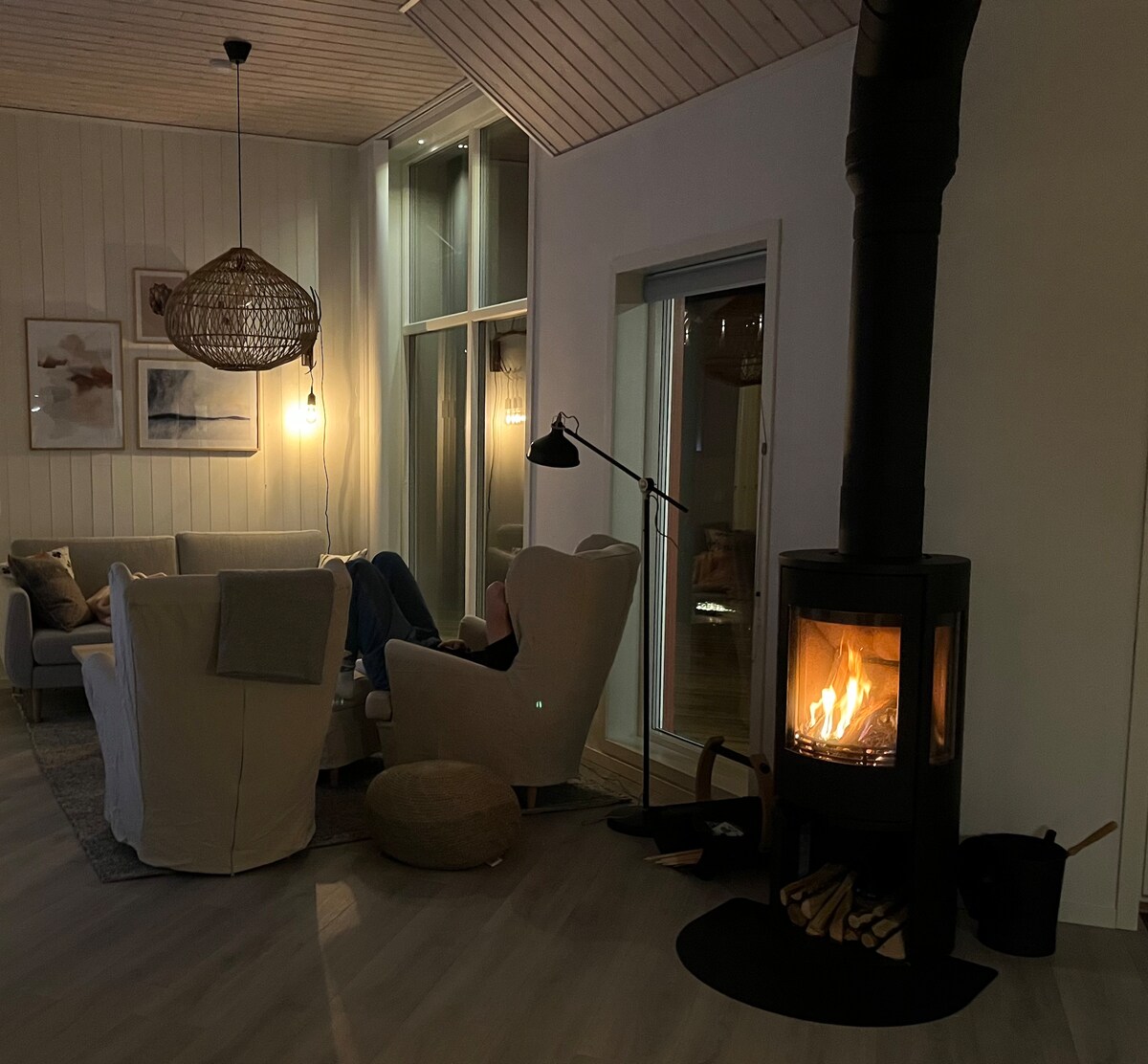 A cozy living area features a modern fireplace with a warm flame, providing a gentle glow. Plush seating arrangements, including a white armchair and comfortable sofa, are complemented by a natural fiber ottoman. Large windows allow light in, enhancing the inviting atmosphere.