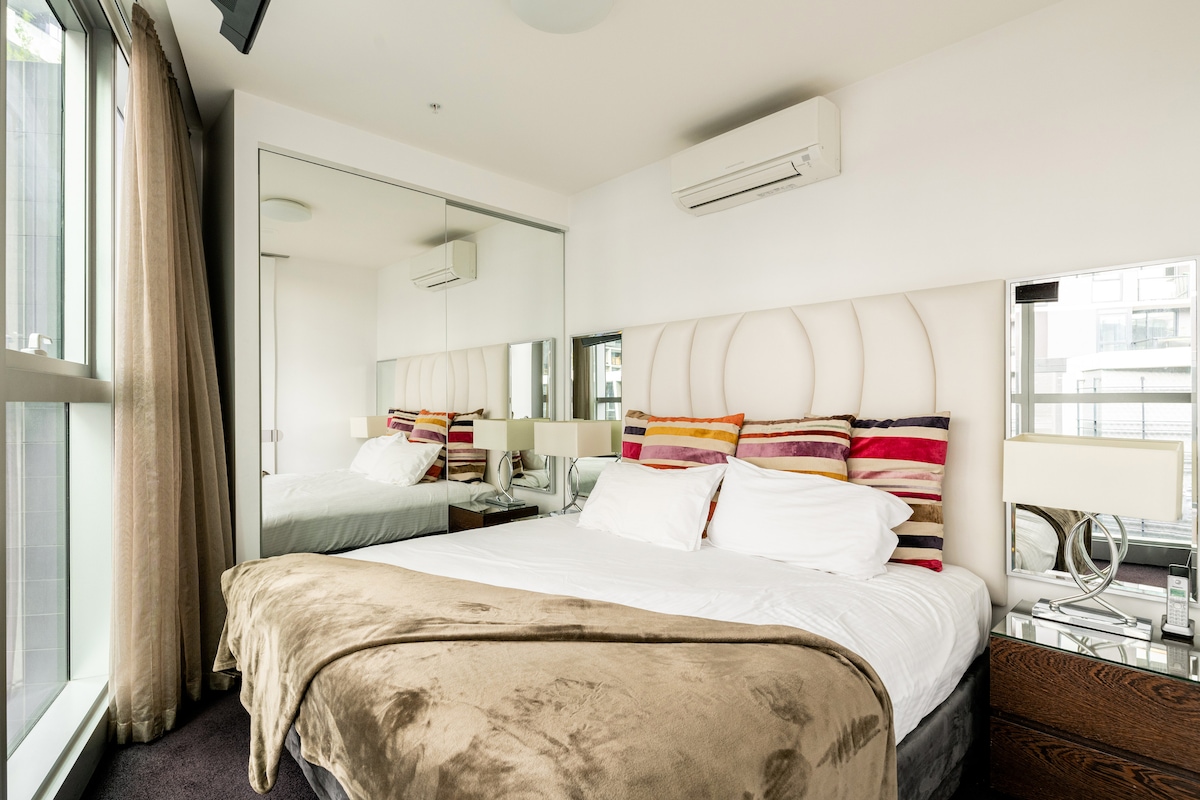 A well-appointed bedroom features a spacious bed with layered linens, accented by vibrant throw pillows. Mirrored panels expand the sense of space while soft natural light filters through large windows, complemented by sheer curtains. Nightstands with lamps provide functional lighting.