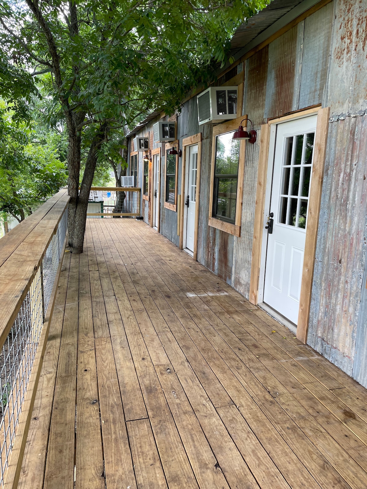 A rustic wooden deck runs alongside a structure featuring corrugated metal walls and large windows. The deck is lined with wooden railings and shaded by trees, providing a natural atmosphere. A few outdoor chairs can be seen in the distance.