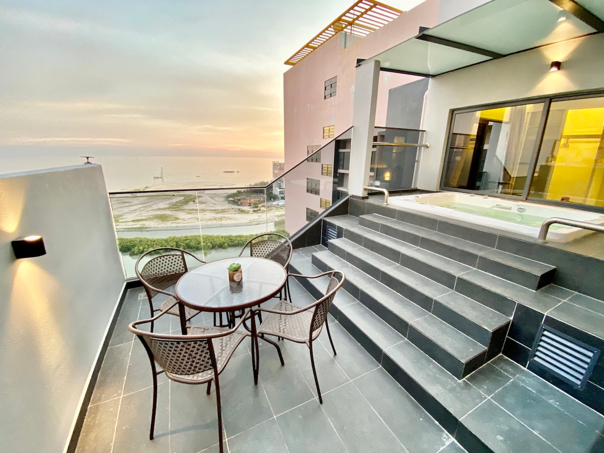 An outdoor terrace area is featured, showcasing a small round table with four chairs. Steps descend to an area with a private jacuzzi, offering views of the sea at sunset. The surrounding architecture includes large windows, illuminating the interior spaces.
