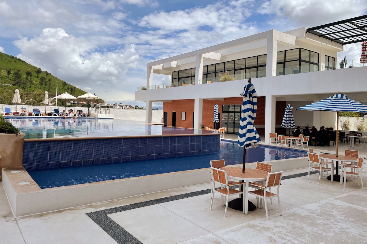 A modern outdoor space features a swimming pool framed by lounge chairs and umbrellas. The building showcases large windows and a spacious terrace, with views of surrounding hills. Tables are arranged nearby, providing a pleasant area for relaxation.