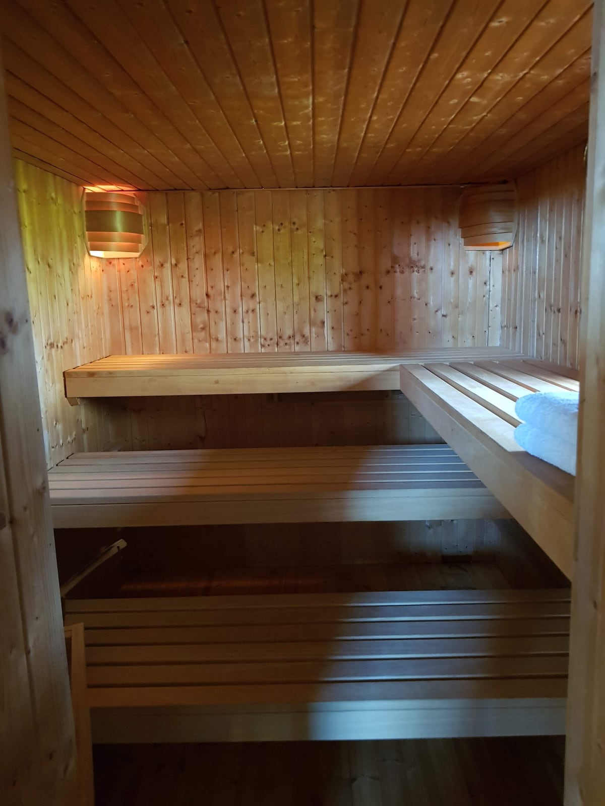 A serene sauna space features wooden benches and walls, providing a warm and inviting atmosphere. Soft lighting from colorful lanterns enhances the calming environment, while neatly rolled towels are placed on one of the benches, inviting relaxation.