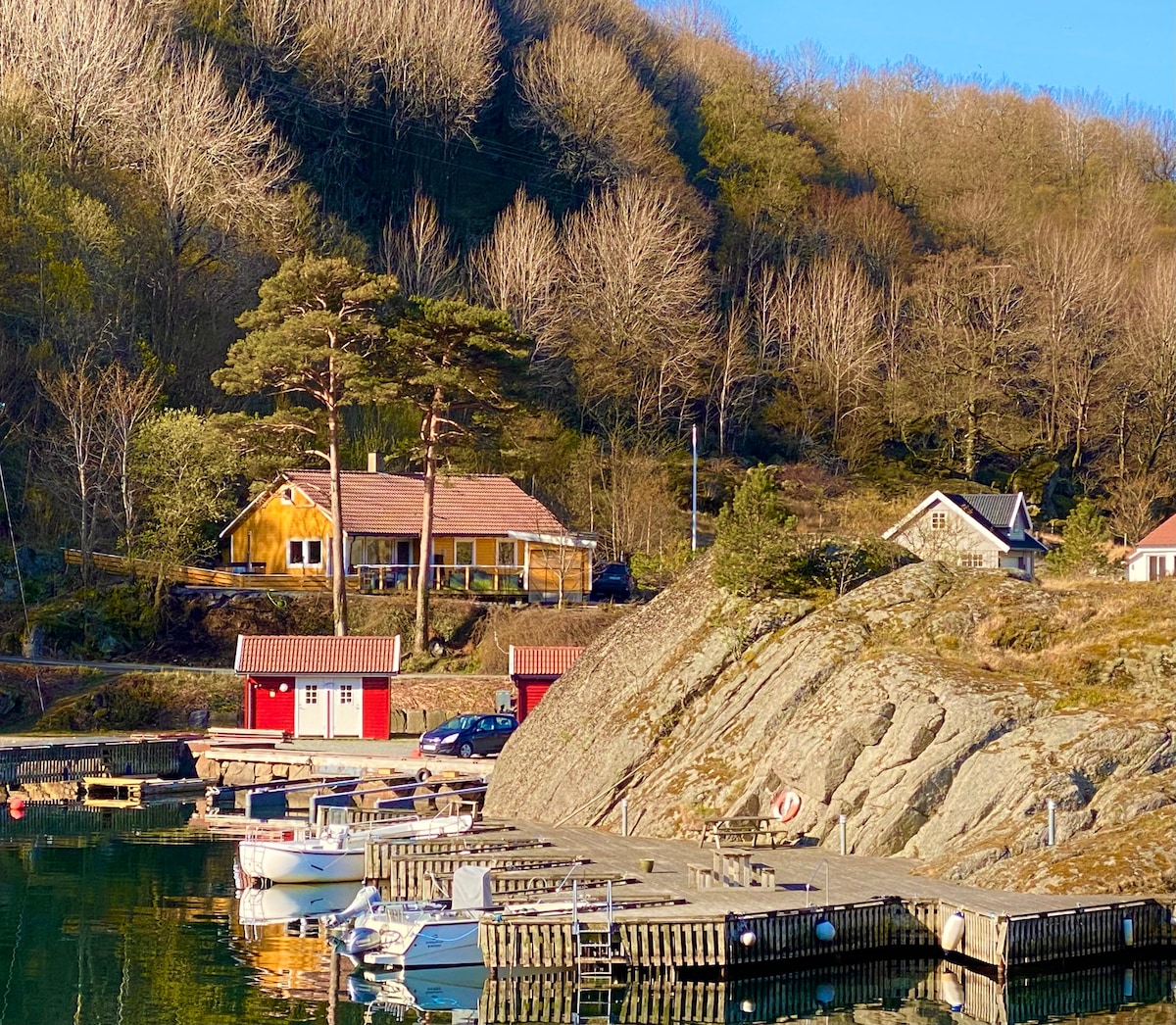 A charming waterside scene captures a collection of boat docks lined with small boats. In the background, a wooden house with a red-roof stands amongst trees, complemented by several nearby cottages. Rocky hills provide a natural backdrop under clear blue skies.