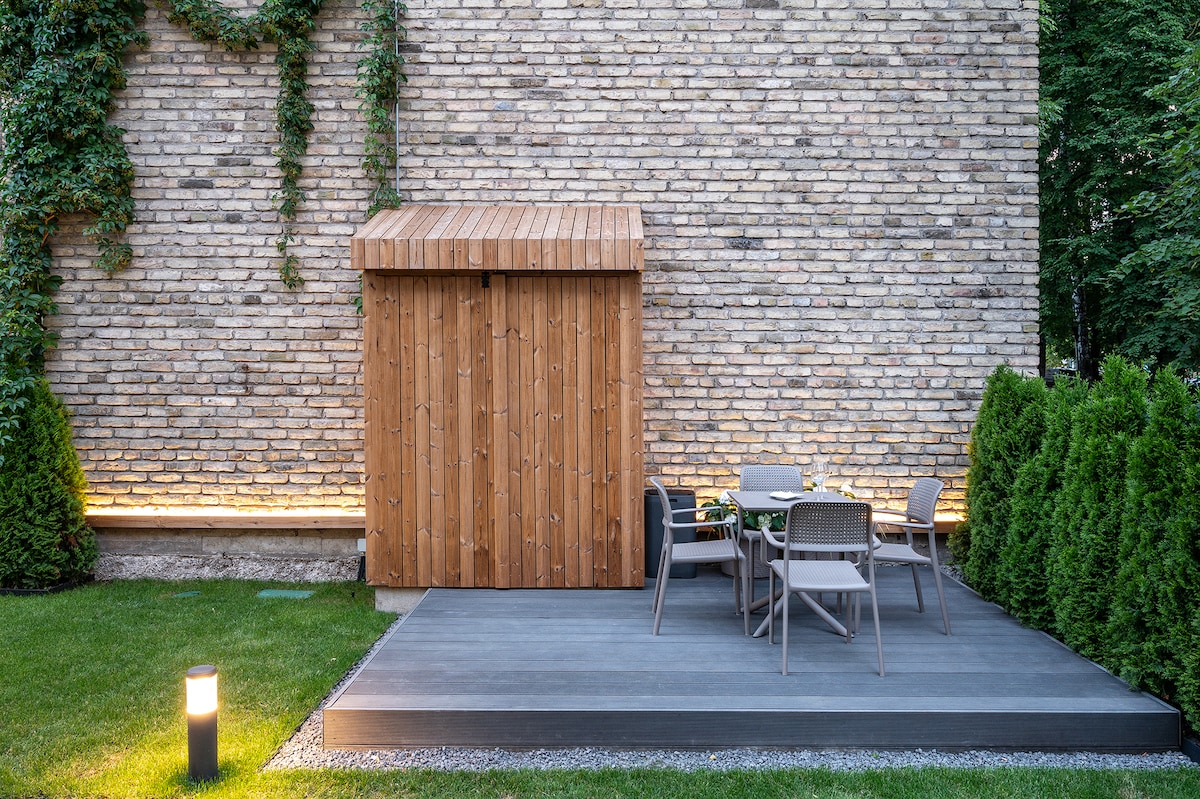 A private backyard features a wooden storage shed against a brick wall, surrounded by lush greenery. A deck area includes a dining table with four chairs, illuminated by soft lighting along the edges. The space provides a calm outdoor setting.